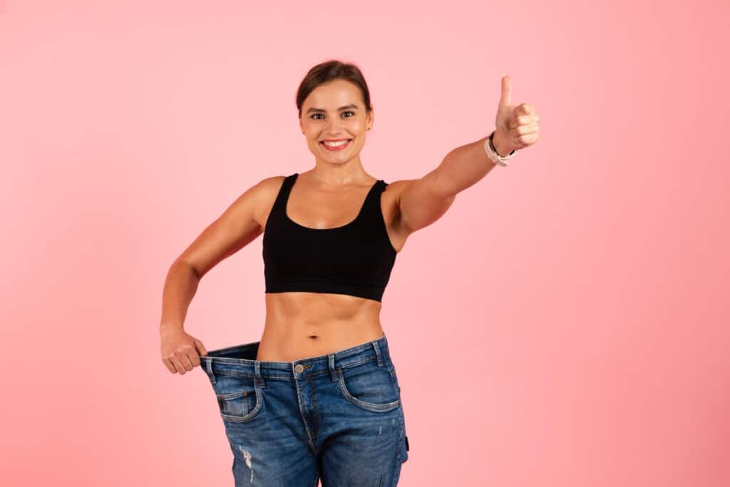 Mosolygós nő fekete topban thumbs up pózban tartja a nagyobb kék farmers nadrágot a derekán, rózsaszín háttérrel – tirzepatid hatóanyag fogyás motivációja.